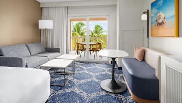 A hotel room with a sofa, a small table, a coffee table, a balcony with chairs, and a painting on the wall.