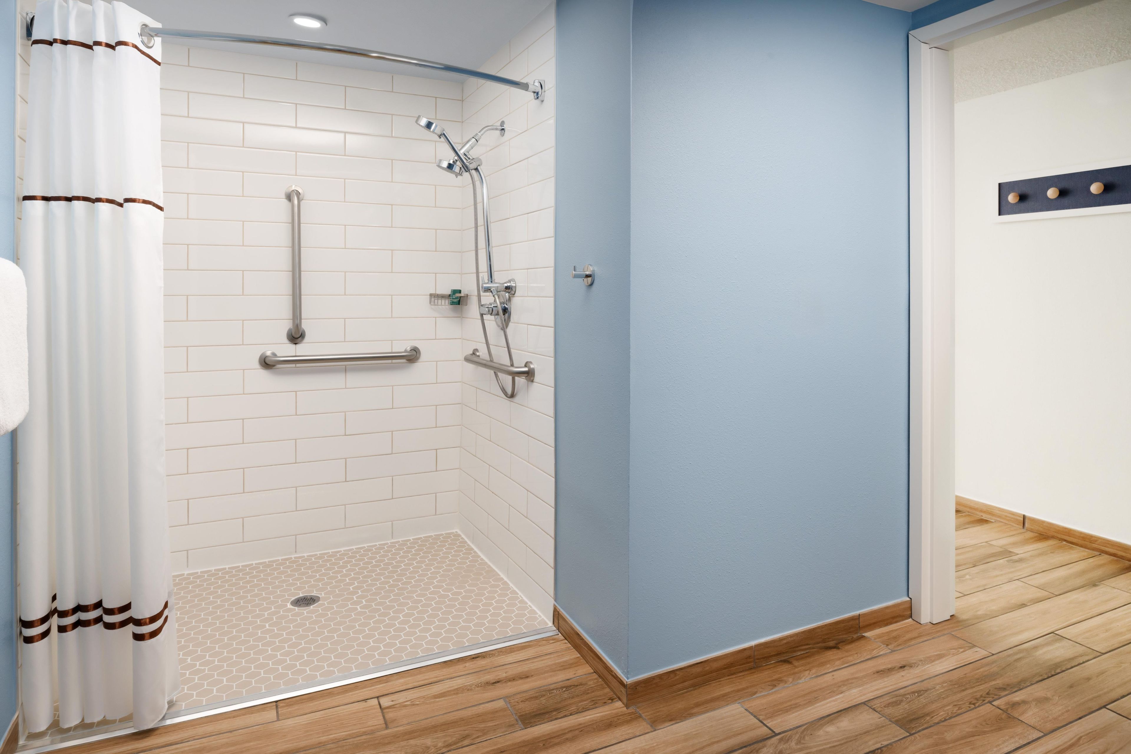A modern bathroom with white tiled walls, a shower with a handheld showerhead, and a wooden floor.