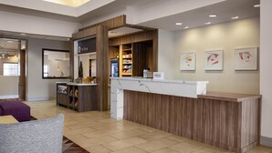 A hotel reception area with a wooden reception desk, a minibar, a seating area with a purple sofa, and framed artwork on the wall. - Hilton Garden Inn Phoenix Airport (Phoenix)