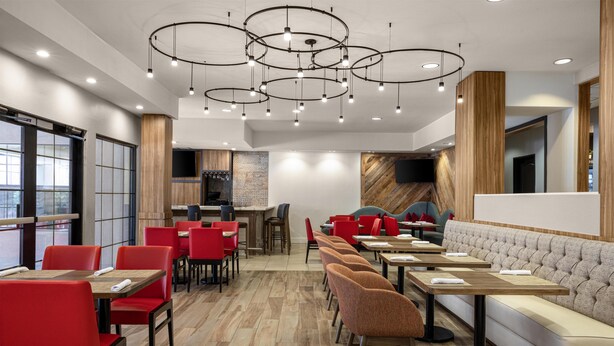 A modern restaurant interior with red and brown chairs, wooden tables, a cushioned bench, and a central bar area. - Hilton Garden Inn Phoenix Airport (Phoenix)