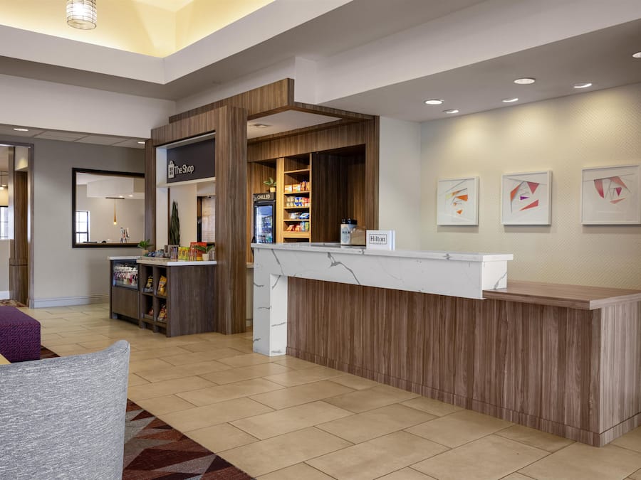 Un espace d’accueil d’hôtel avec un comptoir de réception en bois, un réfrigérateur pour boissons fraîches et un présentoir avec des en-cas. Des œuvres d’art encadrées ornent le mur, et un coin salon est agrémenté d’un tapis à motifs.