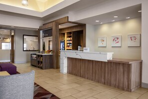 A hotel reception area with a wooden reception desk, a chilled beverage refrigerator, and a counter with snacks. There are framed artworks on the wall and a seating area with a patterned rug. - Hilton Garden Inn Phoenix Airport (Phoenix)