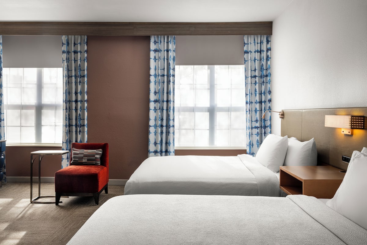 A hotel room with two beds, a red chair, a small table, and a lamp.