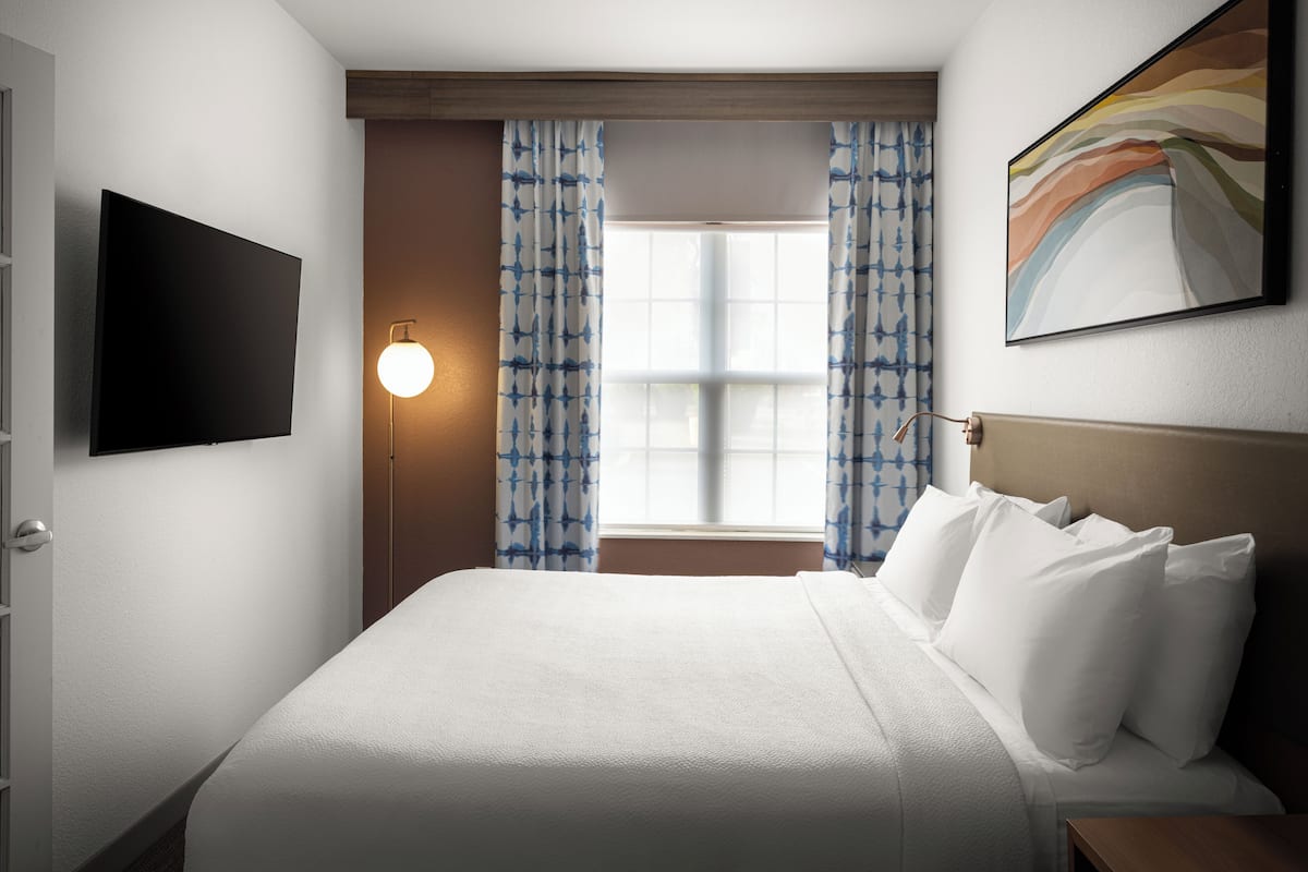 A hotel room with a bed, a flat-screen TV, a wall-mounted lamp, and a window with curtains.