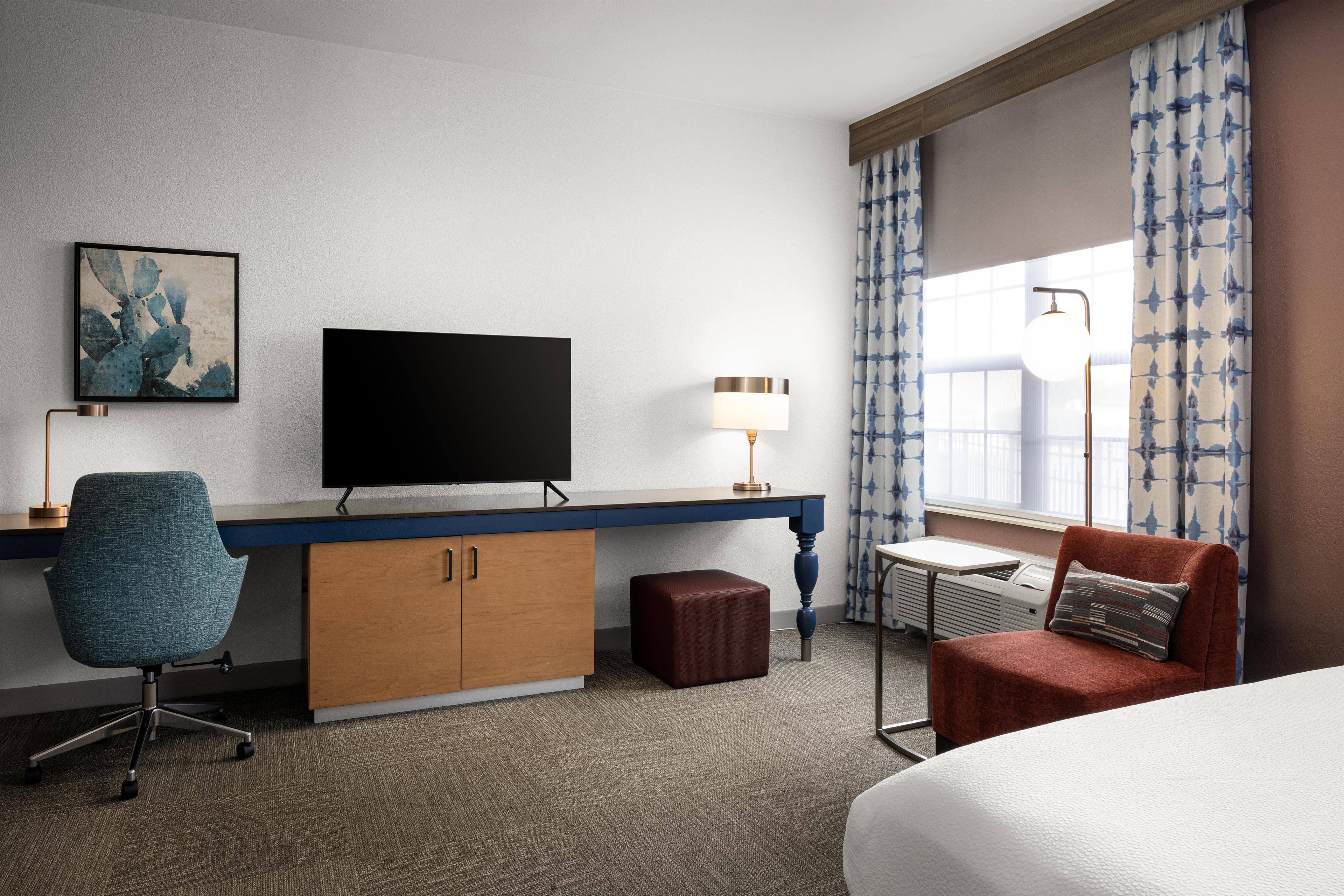 A hotel room with a flat-screen TV, a desk, a chair, a bed, a small table, a lamp, and a window with curtains.