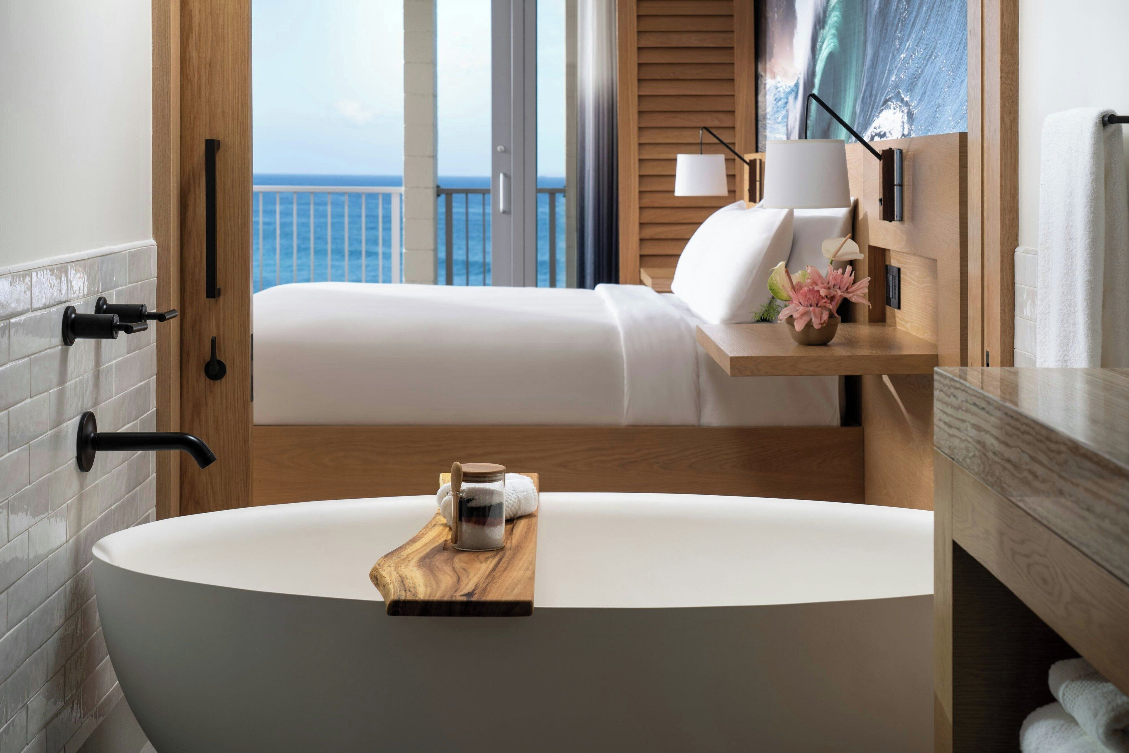 Une salle de bain moderne avec une baignoire sur pieds, un meuble-lavabo en bois, une table de chevet avec une lampe, et une vue sur l’océan par une porte ouverte.
