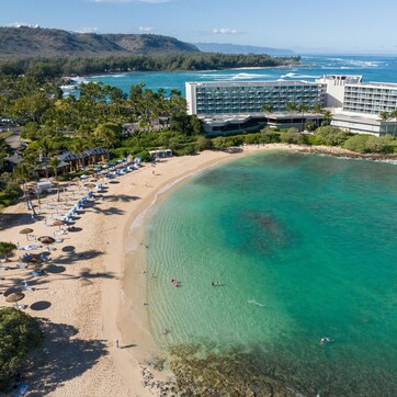 An aerial view of a beach resort with a large building, clear turquoise waters, and a sandy beach with umbrellas.
