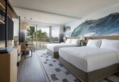 A hotel room with two beds, a desk, and a balcony with a view of palm trees.