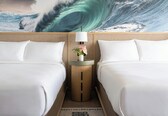 A hotel room with two beds, a wooden side table with a lamp, and a large wave-themed wall mural.