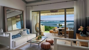 A modern living room with a sofa, coffee table, and ottomans, a dining area with a table and chairs, and a large sliding glass door leading to a balcony with a view of the ocean.