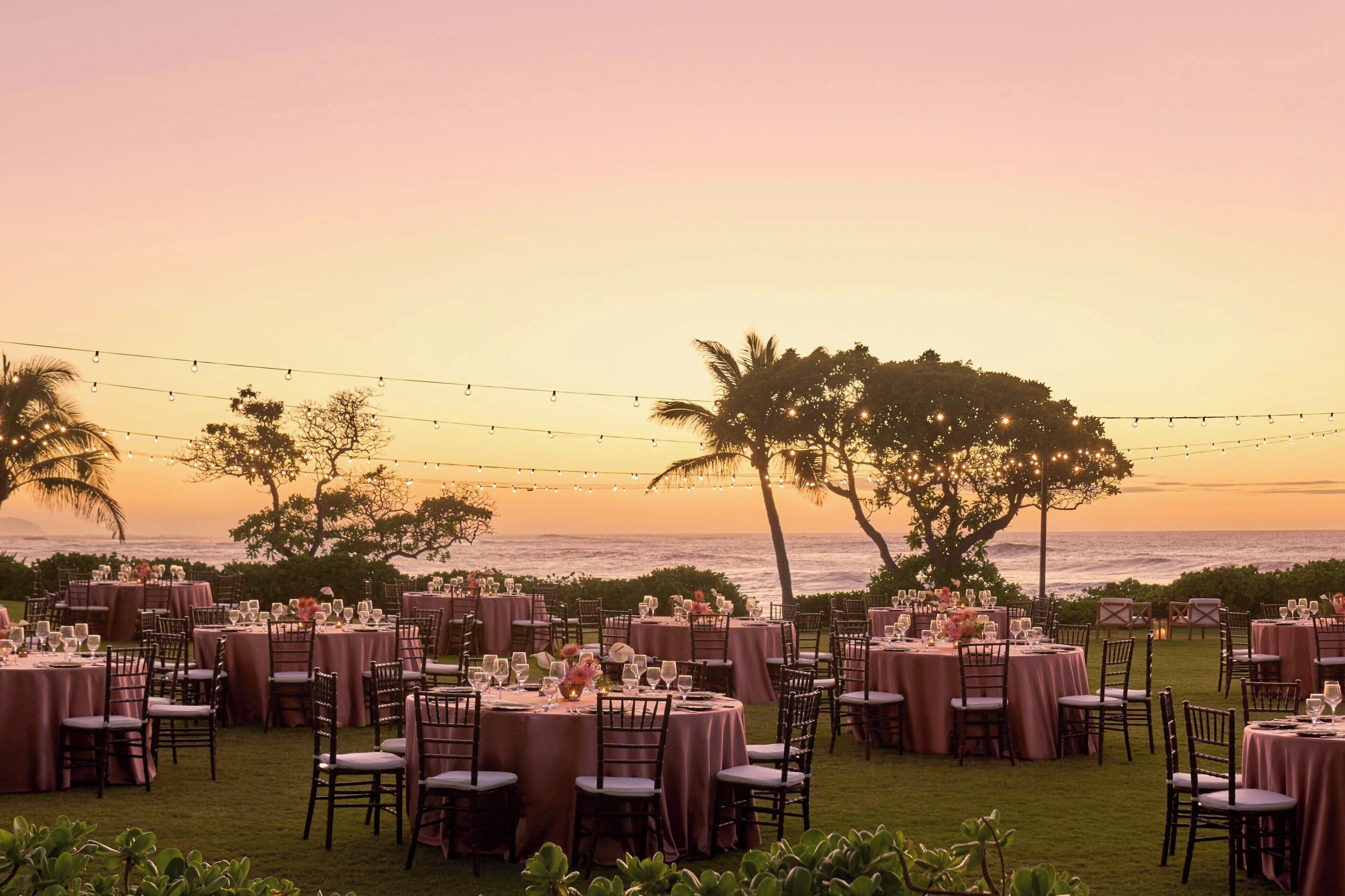An outdoor dining area with round tables and chairs, set against a backdrop of a sunset over the ocean.