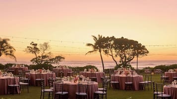 An outdoor dining area with round tables and chairs, set against a backdrop of a sunset over the ocean.