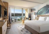 A modern hotel room with a large bed, a desk, and a view of the ocean through a sliding door.