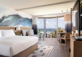 A hotel room with a large bed, a view of the ocean, a dining area with a table and chairs, and a balcony with a view of the sea.