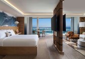 A modern hotel room with a large bed, a TV, a balcony with a view of the ocean, and a seating area with a leather chair and ottoman.