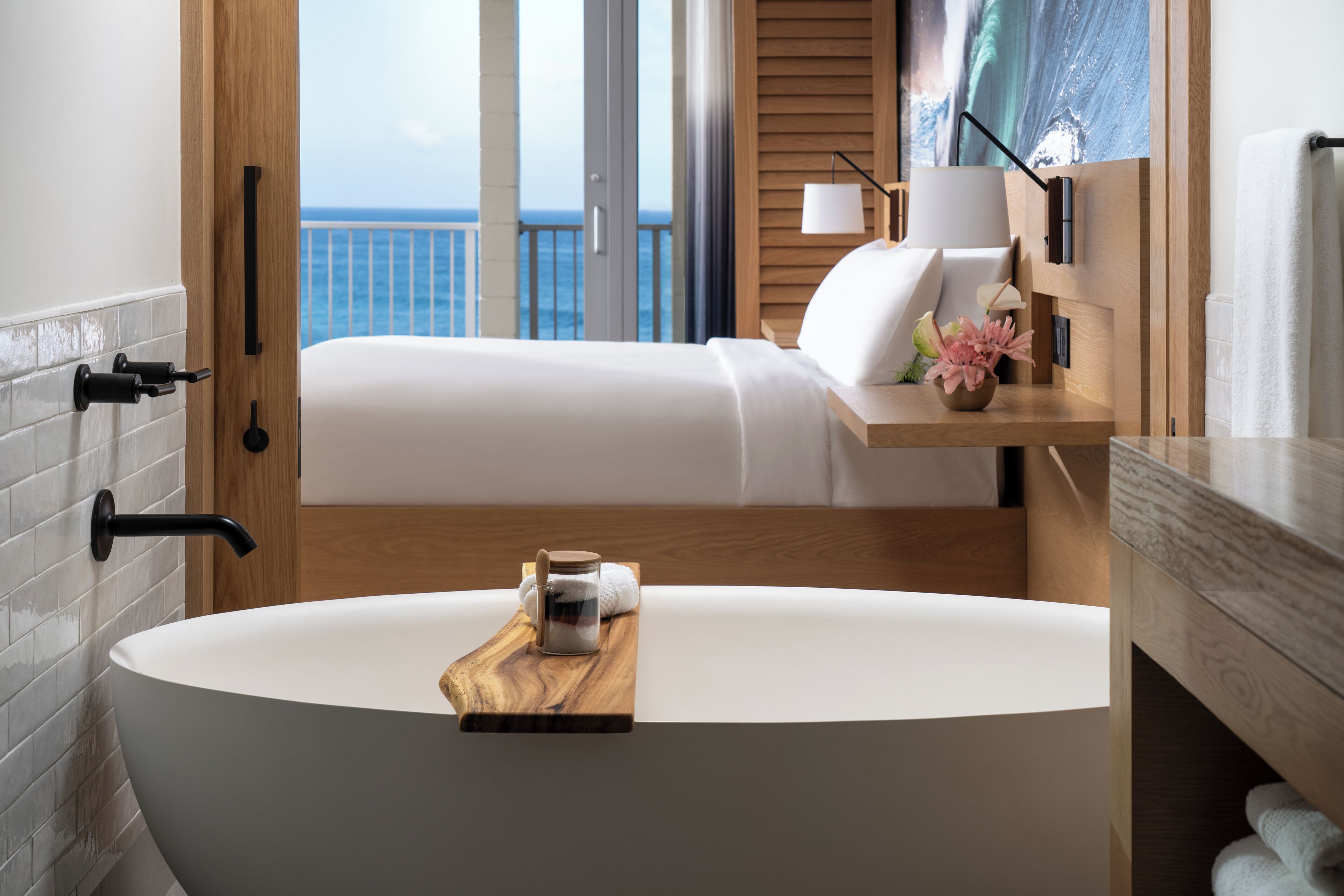 A modern bathroom with a freestanding bathtub, a wooden tray with a jar, and a view of the ocean through a sliding door.