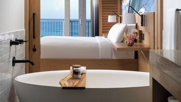 A modern bathroom with a freestanding bathtub, a wooden tray with a jar, and a view of the ocean through a sliding door.