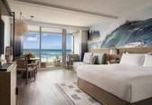 A hotel room with a large bed, a desk, a chair, a small table with a vase, and a view of the ocean through a sliding door.