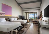 A modern hotel room with two beds, a desk, a chair, and a large window with a view of the ocean.