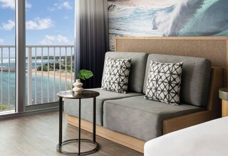 A hotel room with a balcony, a sofa with patterned cushions, a small round table with a vase, and a large ocean mural on the wall.