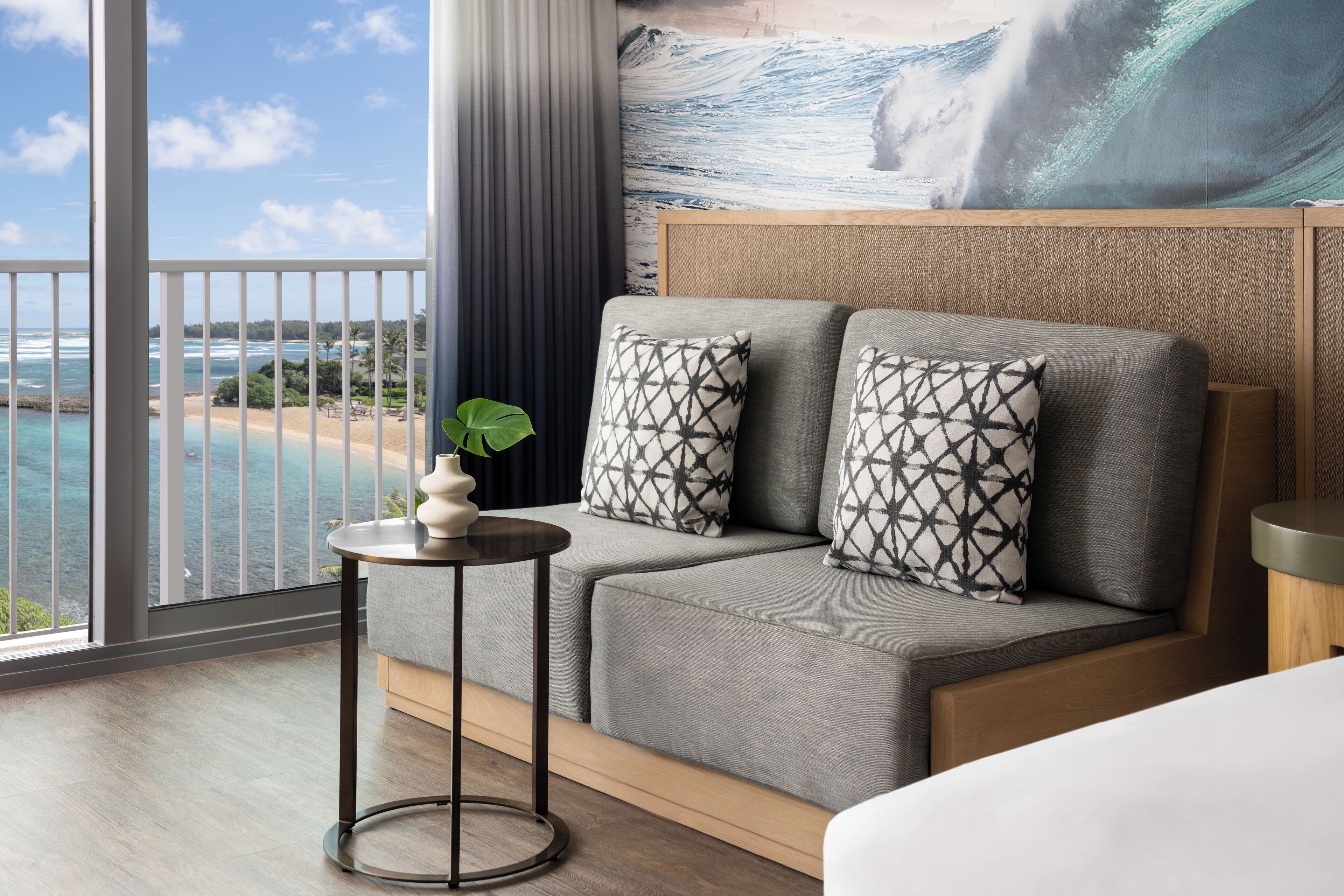 Habitación de hotel moderna con balcón, amplia vista al océano, sofá con cojines estampados y una mesita auxiliar con un florero.