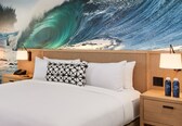 A hotel room with a large wave mural on the wall, a bed with white linens, two bedside lamps, a nightstand with a phone, and two water bottles on the nightstand.