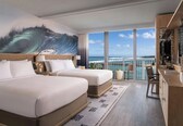 A hotel room with two beds, a large ocean wave mural, a balcony with a view of the ocean and a pier, and a desk with a chair.