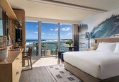 A hotel room with a large bed, a desk, a chair, a small table, and a balcony with a view of the ocean.