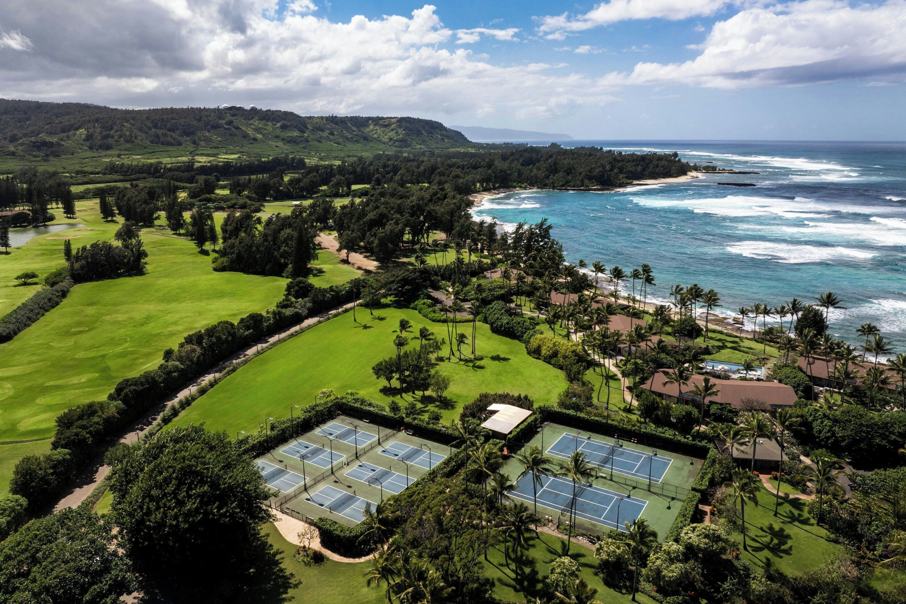 A golf course with multiple tennis courts, surrounded by lush greenery and a view of the ocean.