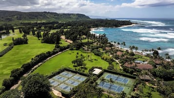 A golf course with multiple tennis courts, surrounded by lush greenery and a view of the ocean.