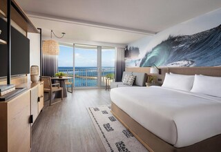 A modern hotel room with a large bed, a flat-screen TV, a dining area with a table and chairs, and a balcony with a view of the ocean.