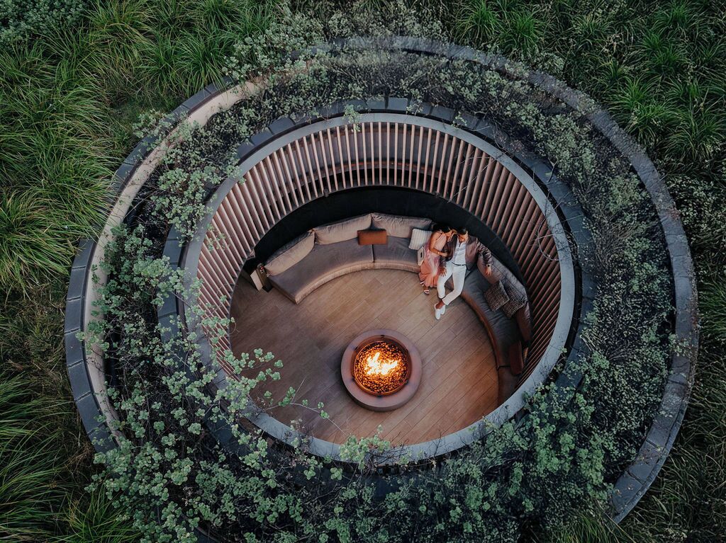Un espacio de estar al aire libre con forma circular, que incluye un brasero en el centro, rodeado por una terraza de madera y un bajo muro de piedra.