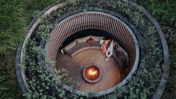 A circular outdoor seating area with a fire pit at the center, surrounded by a wooden deck and a low stone wall.
