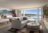 A modern hotel room with a large bed, a sofa, a round ottoman, and a balcony with a view of the sea.