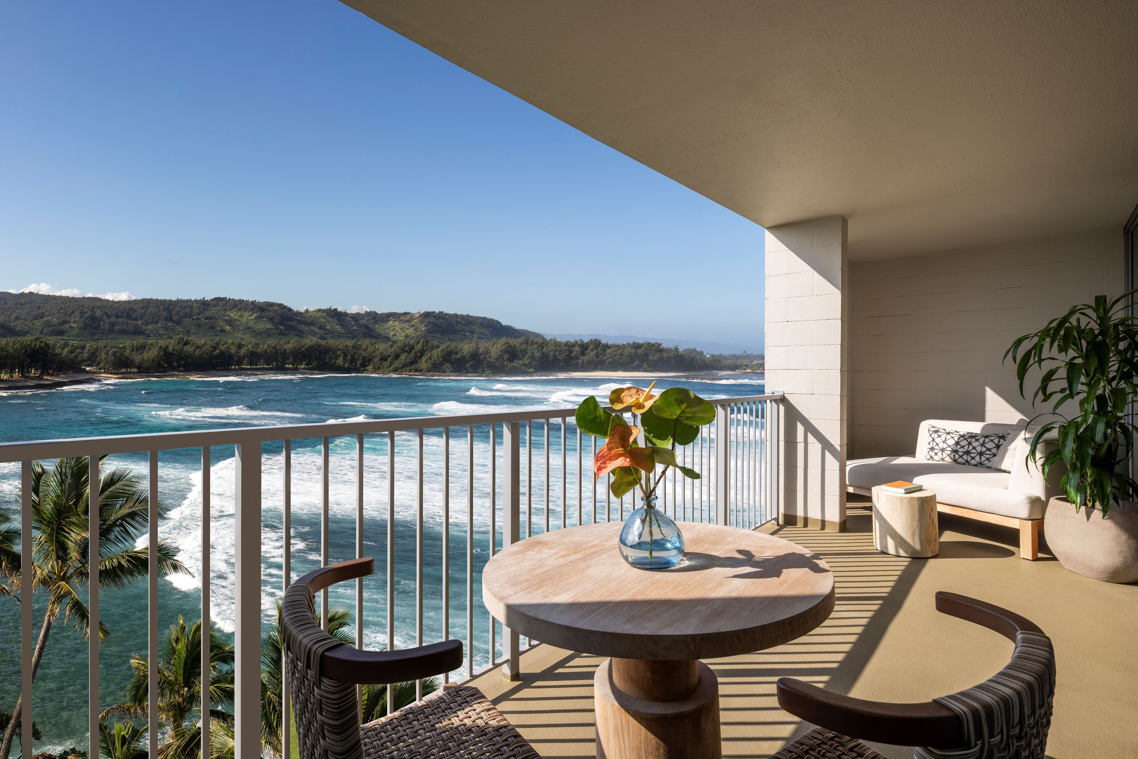A balcony with a view of the ocean, a round wooden table with a vase, wicker chairs, and a sofa.