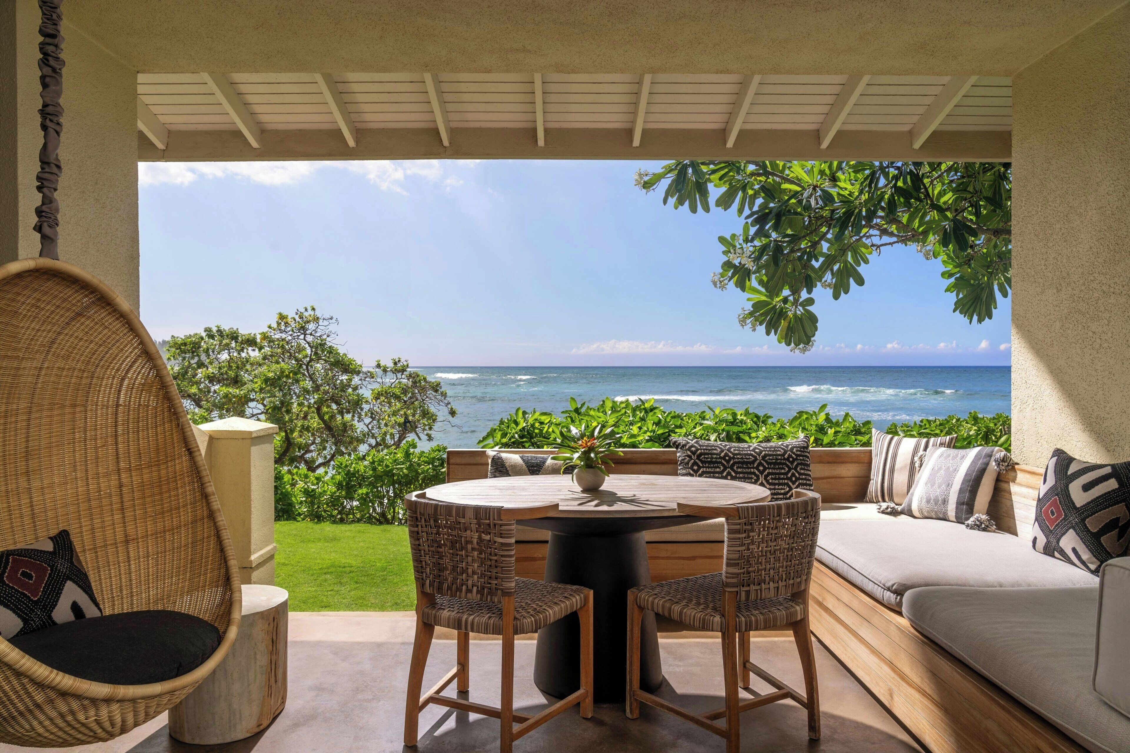 A patio with a round table, two chairs, a sofa, and a hanging chair. The patio overlooks a view of the ocean.