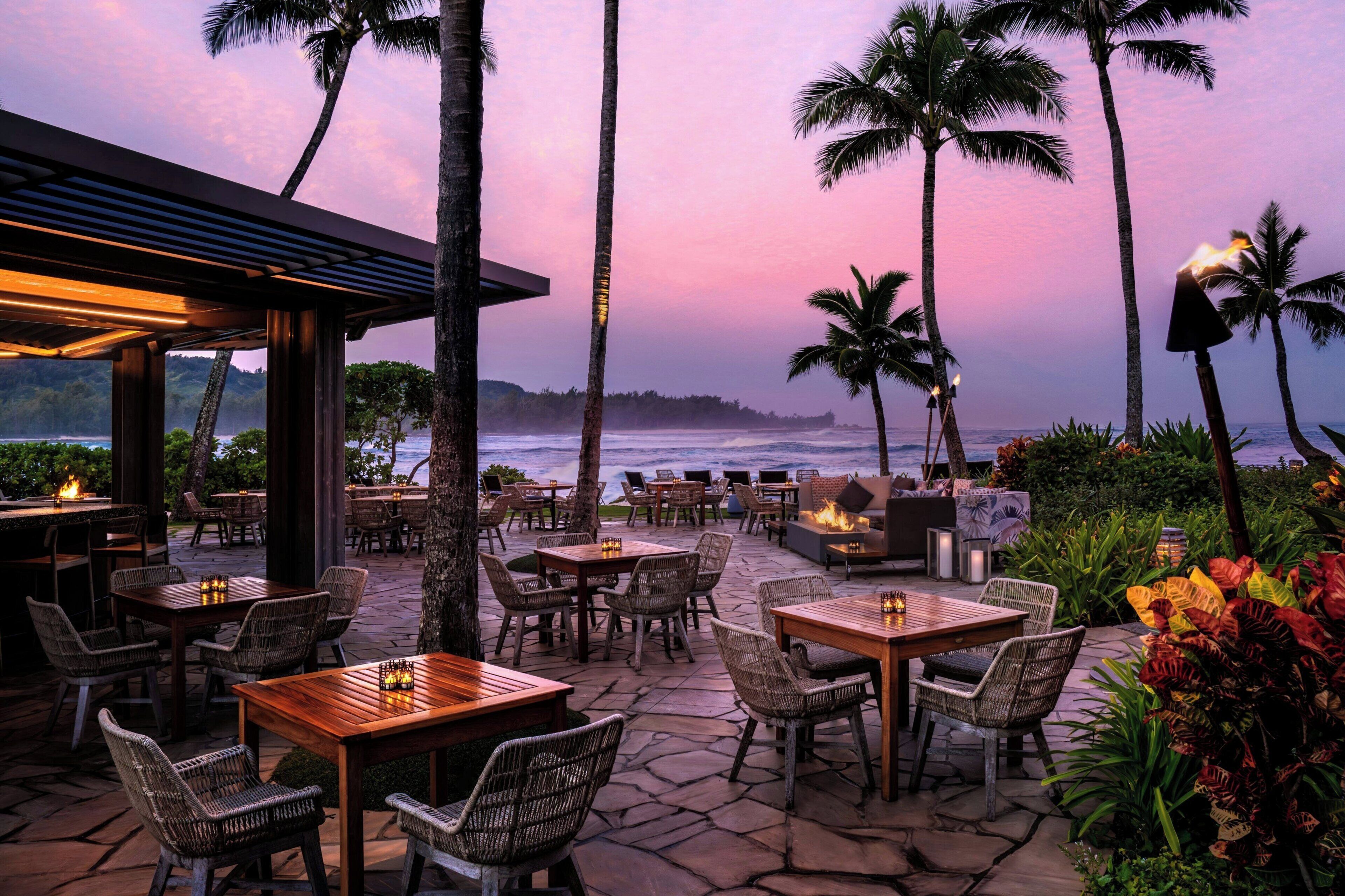 An outdoor dining area with tables and chairs, surrounded by palm trees and a view of the ocean at sunset.