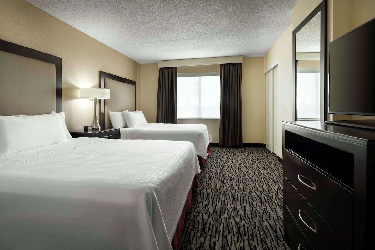 A hotel room with two beds, a desk, a dresser, a television, and a window with curtains.