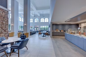 A spacious indoor area with high ceilings, large windows, and a central dining area with tables and chairs.