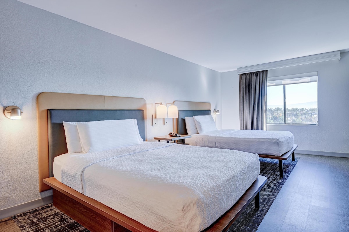 A hotel room with two beds, a large window with curtains, and a view of the city.