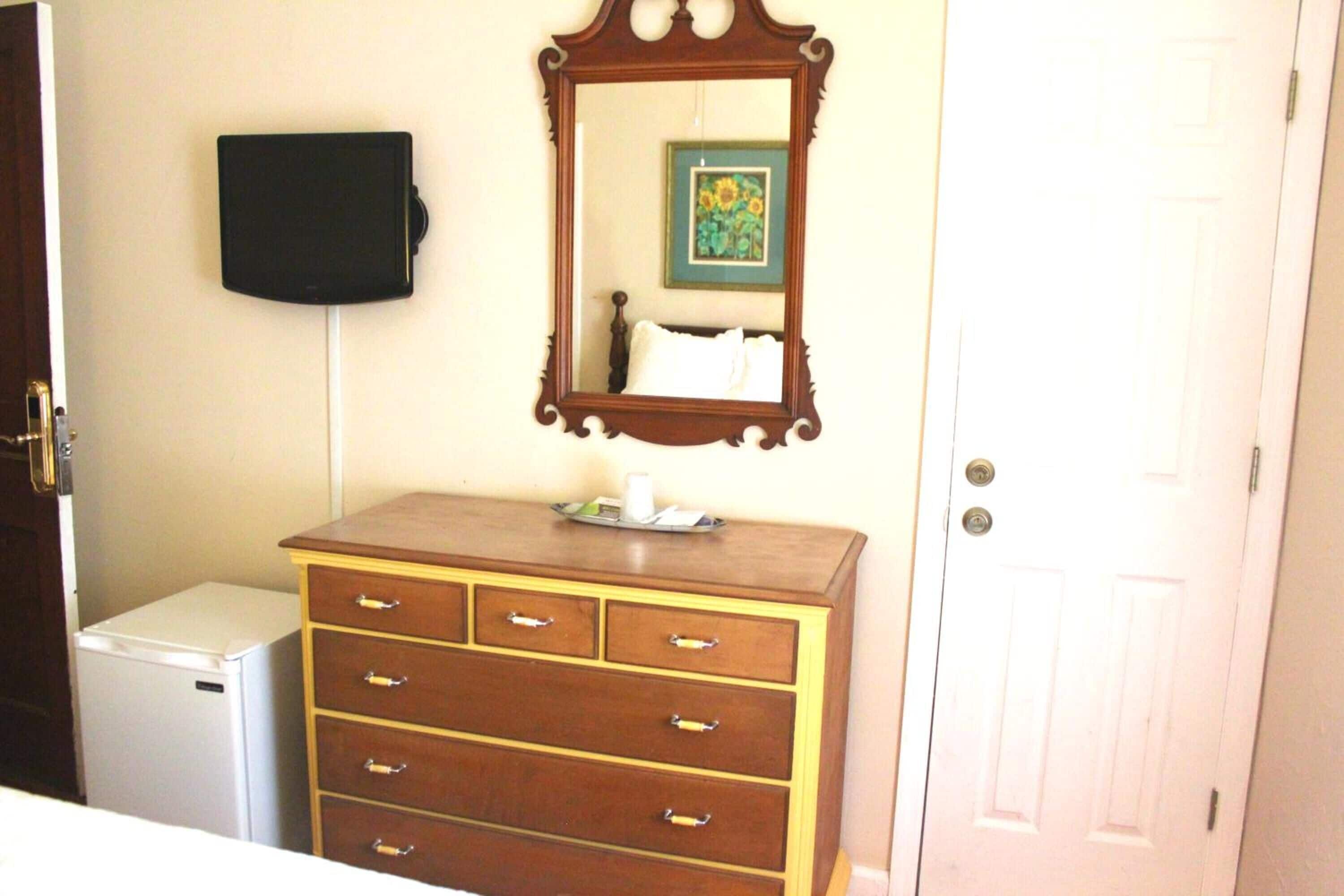 A room with a wooden dresser, a small white refrigerator, a mirror, and a door.