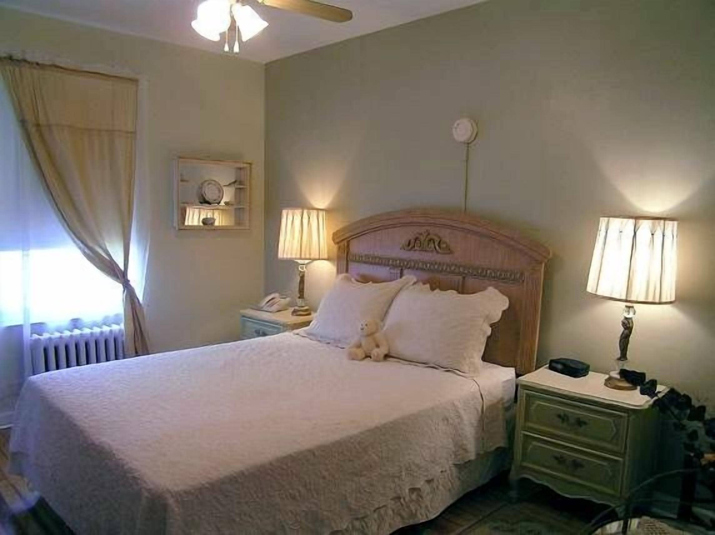 Una cama bien hecha con cabecera, dos lámparas de noche, una mesita de noche y un reloj de pared.