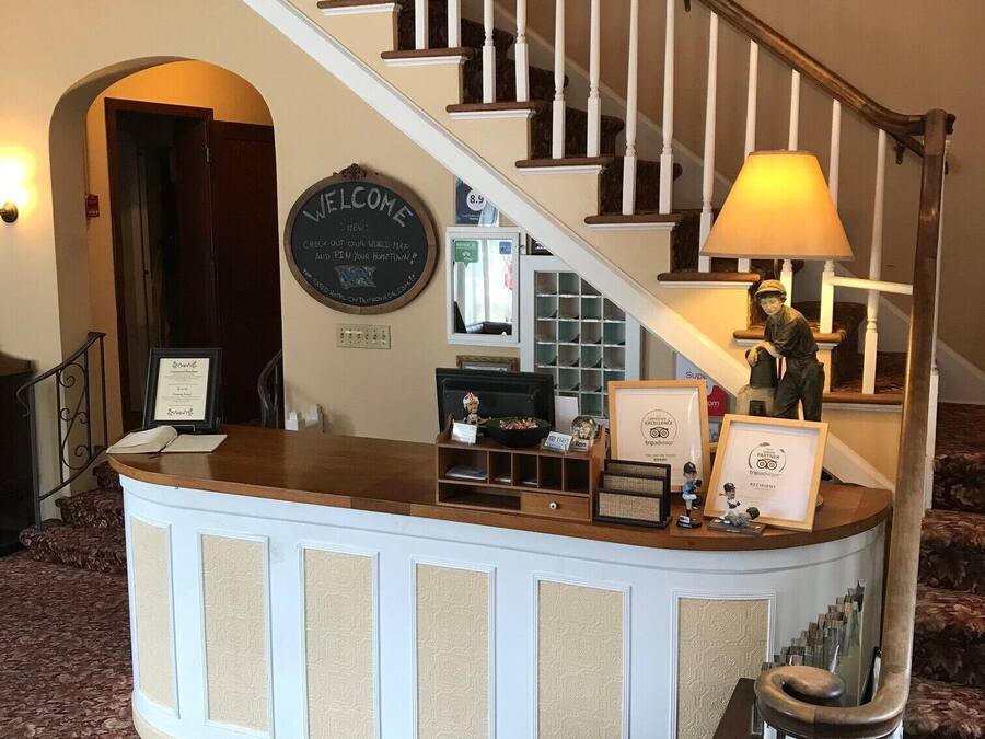 A reception desk with a 'Welcome' sign, a framed certificate, and a lamp. A staircase with a wooden railing and a lamp is visible in the background.