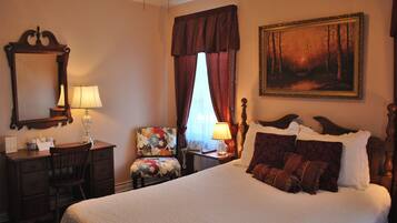 A hotel room with a large bed, a desk with a lamp, a chair, a painting on the wall, and a window with curtains.