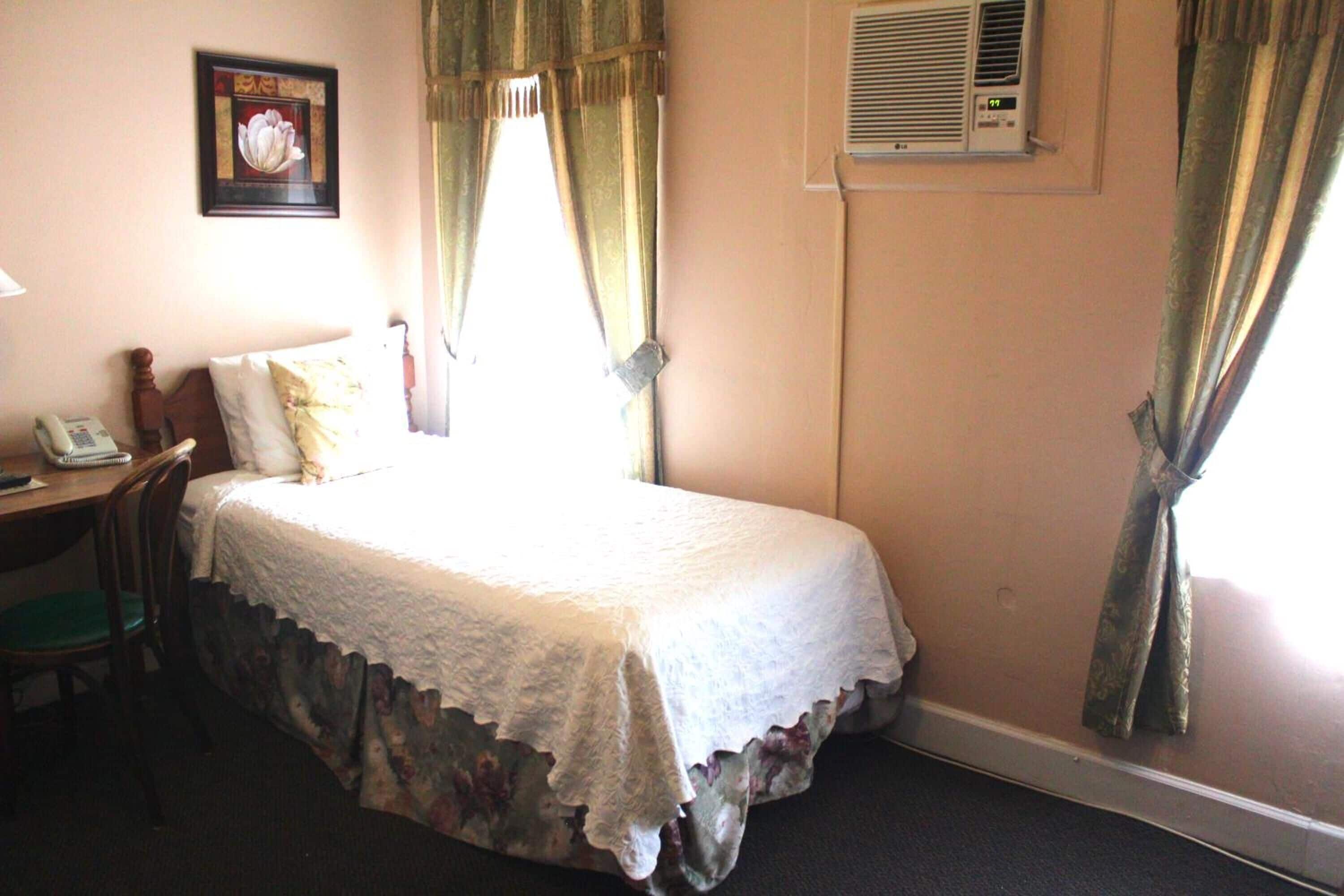 Une chambre d’hôtel avec un lit simple, un bureau avec un téléphone, une chaise et un climatiseur fixé au mur.
