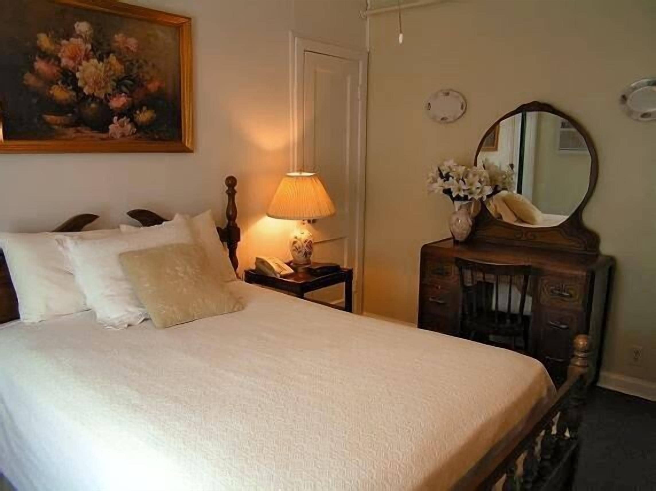 Un lit bien fait, avec une couette blanche et des oreillers, une table de chevet avec une lampe et un vase de fleurs, et une commode en bois avec un miroir.