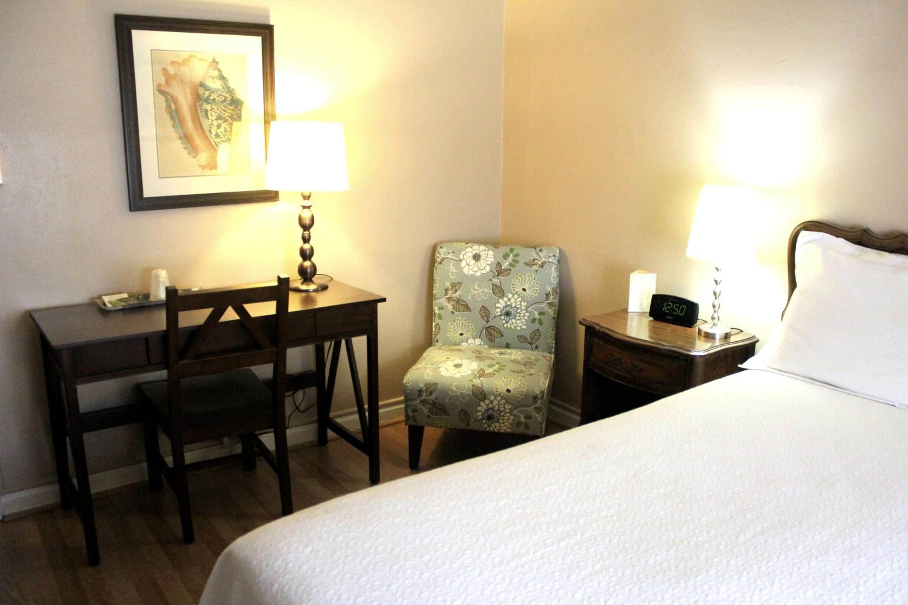 A hotel room with a bed, a desk, a chair, two bedside lamps, and a clock on the nightstand.