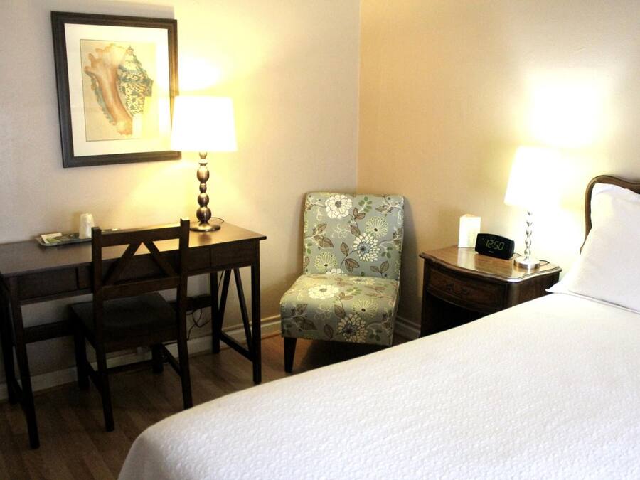 A hotel room with a bed, a desk, a chair, two bedside lamps, and a clock on the nightstand.