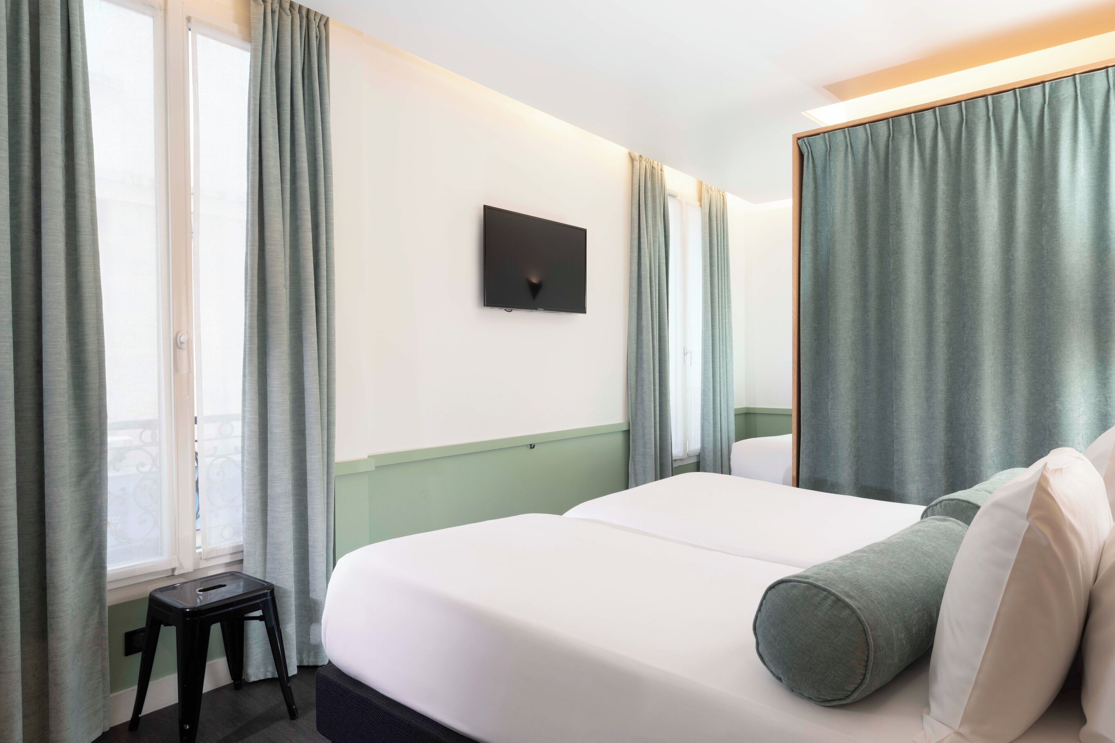 Habitación de hotel con cama, un taburete, televisor de pantalla plana en la pared y cortinas en las ventanas.