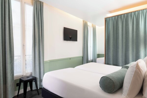 Habitación de hotel con cama, un taburete, televisor de pantalla plana en la pared y cortinas en las ventanas.